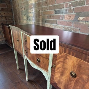 May include: A wooden sideboard featuring a dark brown top surface, two central drawers, and two side cabinets with light green painted legs and trim. The furniture includes circular brass hardware and the word Sold printed in black on a white square.