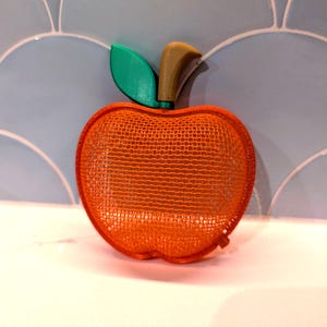 May include: An apple-shaped object constructed from an orange mesh material. The top features a solid green leaf and a brown stem. The structure is hollow and rounded, mimicking the form of a piece of fruit with a distinct stem and leaf attachment.