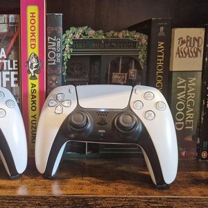 May include: Two white and black wireless video game controllers featuring dual analogue sticks, a directional pad, and four action buttons. The controllers have a smooth, ergonomic design with a central touchpad and light bars visible on the top edges.