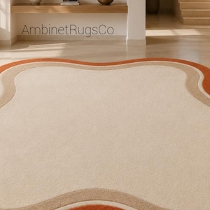 Op de afbeelding: Een vloerkleed met een onregelmatige, golvende, wolkachtige vorm met een groot cr&egrave;mekleurig midden, omringd door concentrische banden van lichtbeige en gebrand oranje. De tekst AmbinetRugsCo is zichtbaar in het bovenste deel van de afbeelding.