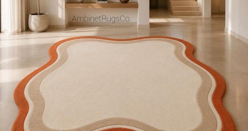 Op de afbeelding: Een vloerkleed met een onregelmatige, golvende, wolkachtige vorm met een groot cr&egrave;mekleurig midden, omringd door concentrische banden van lichtbeige en gebrand oranje. De tekst AmbinetRugsCo is zichtbaar in het bovenste deel van de afbeelding.