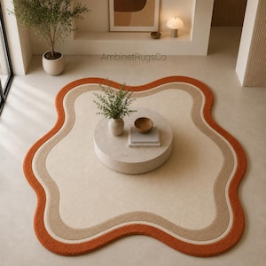Op de afbeelding: Een onregelmatig, wolkvormig vloerkleed met concentrische golvende banen in cr&egrave;me, beige en oranje. Het kleed ligt in het midden van een vloer met een ronde salontafel waarop een kleine plant en een stapel boeken zijn geplaatst.