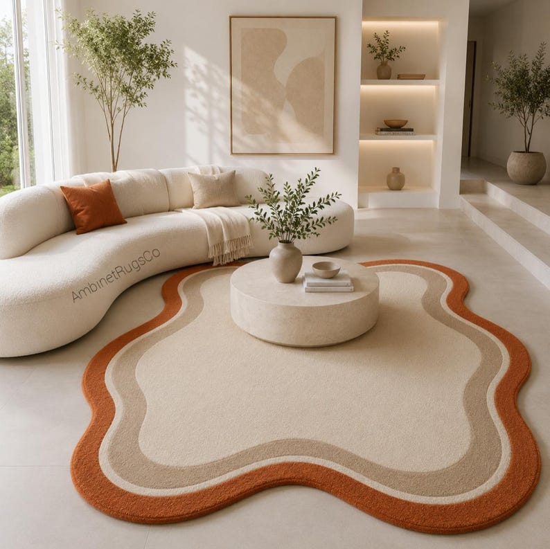Op de afbeelding: Een onregelmatig gevormd vloerkleed met organische, concentrische golvende banen in cr&egrave;me, beige en gebrand oranje. Het kleed ligt op een lichte vloer voor een gebogen cr&egrave;mekleurige bank met een kussen in gebrand oranje.