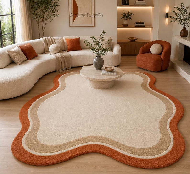 Op de afbeelding: Een vloerkleed met een onregelmatige, organische vorm met een groot cr&egrave;mekleurig midden, omringd door concentrische golvende banden in lichtbruin en gebrand oranje. Het kleed heeft een getextureerd oppervlak en ligt op een lichte houten vloer.