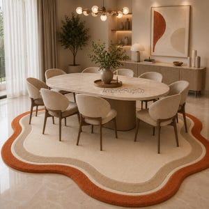 Op de afbeelding: Een onregelmatig, golvend vloerkleed met concentrische banen in cr&egrave;me, beige en gebrand oranje. Het kleed ligt onder een ovale eettafel met een licht stenen blad en een cilindrische voet, omringd door acht gestoffeerde stoelen.