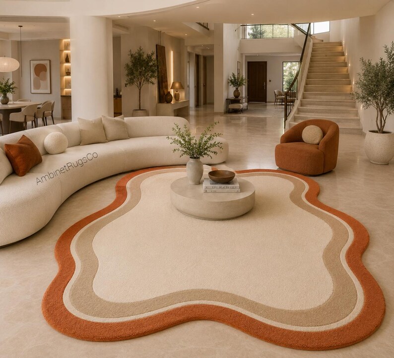 Op de afbeelding: Een vloerkleed met een onregelmatige, organische vorm, voorzien van een cr&egrave;mekleurig midden omringd door concentrische golvende banden in lichtbeige en gebrand oranje. Het kleed ligt op een lichte vloer in een kamer met een gebogen bank.