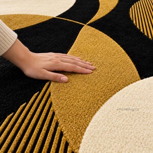 Op de afbeelding: Een pluche vloerkleed met een geometrisch patroon van grote zwarte, mosterdgele en cr&egrave;mekleurige halve cirkels en cirkels. Sommige delen vertonen een gestructureerd, geribbeld lineair ontwerp in mosterdgeel en zwart. De tekst AmbinetRugCo is zichtbaar.