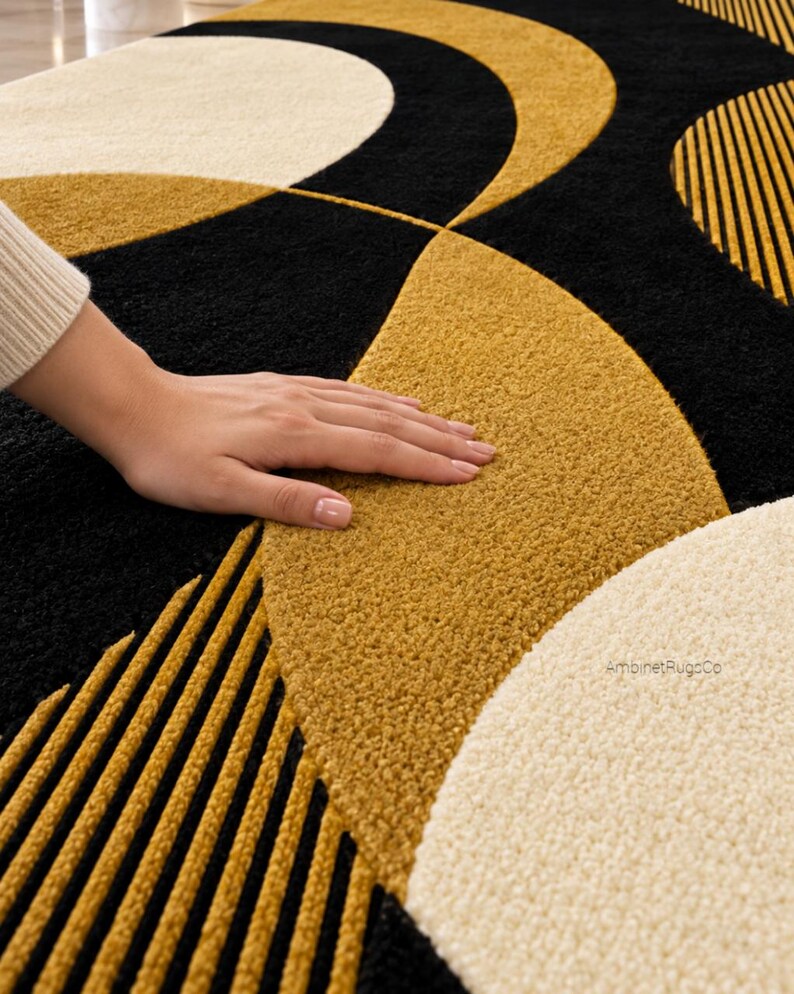 Op de afbeelding: Een pluche vloerkleed met een geometrisch patroon van grote zwarte, mosterdgele en cr&egrave;mekleurige halve cirkels en cirkels. Sommige delen vertonen een gestructureerd, geribbeld lineair ontwerp in mosterdgeel en zwart. De tekst AmbinetRugCo is zichtbaar.