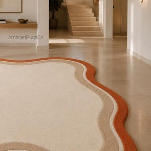 Op de afbeelding: Een vloerkleed met een onregelmatige, golvende vorm met een groot cr&egrave;mekleurig midden, omrand door een dunne witte lijn, een bredere beige band en een dikke buitenste oranje rand. De tekst AmbinetRugsCo is zichtbaar op de muur boven het kleed.