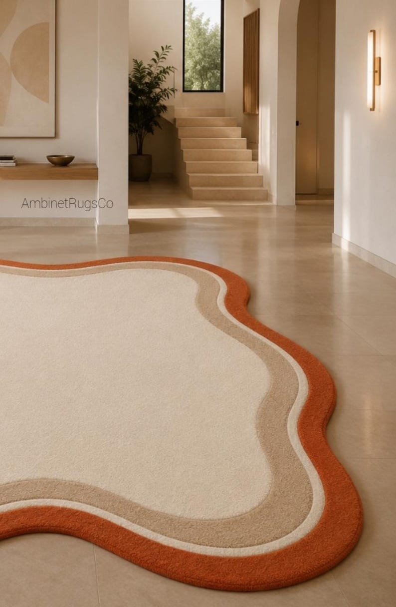 Op de afbeelding: Een vloerkleed met een onregelmatige, golvende vorm met een groot cr&egrave;mekleurig midden, omrand door een dunne witte lijn, een bredere beige band en een dikke buitenste oranje rand. De tekst AmbinetRugsCo is zichtbaar op de muur boven het kleed.