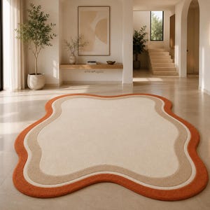Op de afbeelding: Een onregelmatig gevormd vloerkleed met een cr&egrave;mekleurig midden, een beige tussenrand en een feloranje buitenrand. Het kleed heeft een zachte, pluizige textuur en ligt op een gladde, lichte vloer in een heldere kamer.