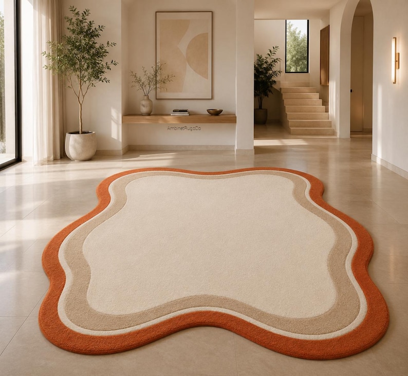 Op de afbeelding: Een onregelmatig gevormd vloerkleed met een cr&egrave;mekleurig midden, een beige tussenrand en een feloranje buitenrand. Het kleed heeft een zachte, pluizige textuur en ligt op een gladde, lichte vloer in een heldere kamer.