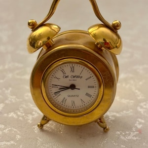 May include: A small, gold-colored quartz alarm clock with a handle and two bells. The clock face is white with black Roman numerals and hands. The clock has three small feet and the text "C.R. WADDOP Est. 1983" is visible on the clock face.