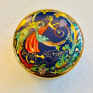 May include: A round metal trinket box with a gold-toned rim and a vibrant enamel lid. The lid features a deep blue background with a central figure in a flowing orange dress, surrounded by intricate green, yellow, and red swirling floral motifs.