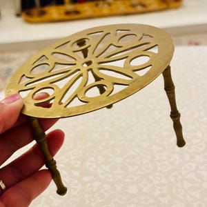 May include: A circular brass trivet featuring an intricate openwork geometric pattern on the top surface. The trivet is supported by three slender, tapered legs that have a segmented, bamboo-like appearance, providing a stable base.