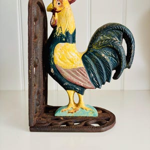 May include: A cast iron bookend featuring a rooster with a yellow neck, dark blue wings and tail, and a red comb. The rooster stands on a light blue base attached to a dark brown, ornate, openwork metal support plate with a weathered finish.