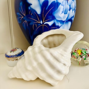 May include: A white ceramic seashell-shaped decorative vase. A blue and white floral vase is in the background, along with a small floral trinket box and a colorful glass paperweight. The seashell vase has a textured surface.