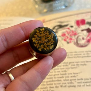 Peut inclure: Un petit bouton rond noir orné d'une fleur jaune séchée et pressée, encapsulée dans une finition en résine transparente et brillante. Le motif floral présente des pétales délicats rayonnant depuis un centre, créant un détail botanique.