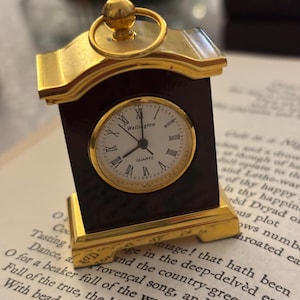 May include: A small, decorative clock with a gold-colored frame and a dark brown base. The clock face is white with black Roman numerals and the word "Wellington" printed on it. The clock is sitting on top of a book.