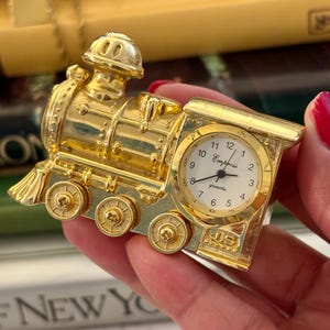 May include: A gold-colored miniature train-shaped quartz clock with a white face. The clock face features black numerals and the text "Empress" and "quartz" in black script. The train body has detailed wheels and a chimney, with "J.S" embossed on the front.