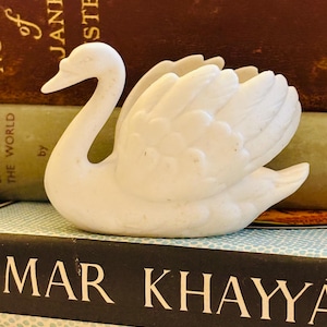 May include: A white swan figurine crafted from a smooth, matte material. The swan is depicted with its neck curved gracefully and its wings partially spread, showing detailed feather textures along the body and wings. The surface is solid white.