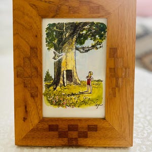May include: A rectangular wooden picture frame with a light brown grain and inlaid geometric square patterns. Inside the frame is a colourful illustration of a person standing before a large tree with an open door, a flying bird, and green grass.