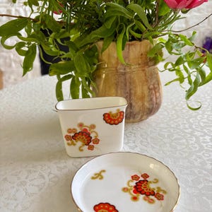 May include: A white porcelain creamer and a matching small round saucer, both featuring a decorative pattern of red and orange fan-shaped motifs with gold accents and thin gold rims. The set is made of smooth, glossy white ceramic material.