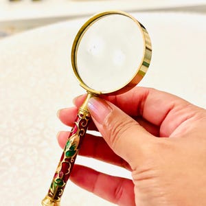 May include: A handheld magnifying glass featuring a circular lens framed in polished gold-tone metal. The cylindrical handle is decorated with an intricate enamel pattern in shades of deep red, green, and gold, ending in a flared base.