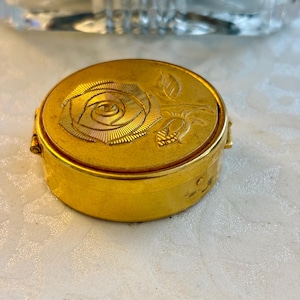 May include: A small, round, gold-colored trinket box. The lid features a detailed embossed rose design. The box is made of metal and has a hinged opening. The box is likely vintage or antique.