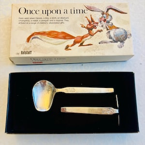 May include: A silver-plated spoon and pusher set in a black box. The box lid features the text Once upon a time and illustrations of a squirrel and rabbit. The spoon bowl has a small star engraving, and the handles feature embossed animal motifs.