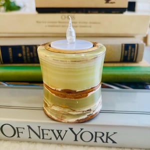 May include: A cylindrical candle holder crafted from banded green and brown stone with a smooth, polished finish. The top features a gold-toned metal rim surrounding a white circular insert with a small, clear, flame-shaped plastic LED light.