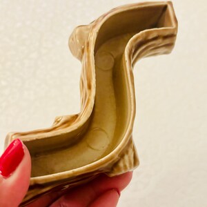 May include: A hollow, S-shaped ceramic object with a light brown, textured exterior that mimics the appearance of tree bark. The interior is smooth and light tan, featuring a curved channel design with two circular indentations at the base.