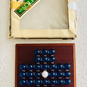 Peut inclure: Un jeu de solitaire House of Marbles avec un plateau en bois carré présentant une grille en forme de croix composée de billes en verre bleu et d'une bille blanche centrale. Le jeu inclut une boîte en carton crème avec une fenêtre transparente.