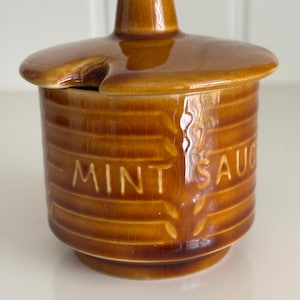 May include: A vintage brown ceramic mint sauce jar with a lid. The jar has a cylindrical shape with horizontal ridges and the words "MINT SAU" are visible. The lid has a small handle and a notch for a spoon.