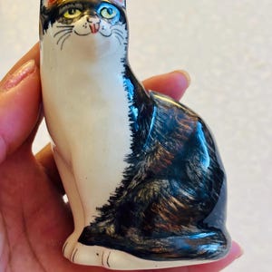 May include: A ceramic cat figurine featuring a glossy finish with a white chest and face, contrasted by black and brown tabby markings on the back and head. The cat has yellow eyes, a small pink nose, and a subtle smile, depicted in a seated position.
