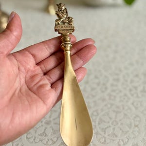May include: A brass shoehorn featuring a decorative handle with a sculpted figure of a pixie sitting on a mushroom. The base of the figure is embossed with the text "CORNISH PIXIE" in capital letters. The metal has a polished, golden finish.