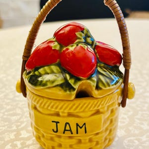 May include: A yellow ceramic jam jar shaped like a woven basket with a lid featuring sculpted red strawberries and green leaves. The jar has a brown, textured handle and the word JAM printed in black letters on the front of the basket base.