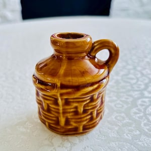 May include: A small, brown, glazed ceramic jug featuring a textured basket-weave pattern on the lower body, a smooth neck, and a single side handle. The glossy finish displays a fine crackle effect throughout the surface of the vessel.