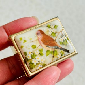 May include: A rectangular metal box with a gold-toned frame featuring an inset panel. The panel displays a painted illustration of a brown bird perched on a branch surrounded by small white flowers with green leaves against a cream background.