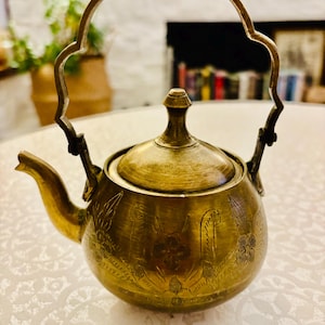 May include: A brass teapot featuring a rounded body with etched floral patterns, a matching lid with a small knob, and a tall, arched handle. The metal has a warm, aged patina, and the spout extends from the side of the vessel.