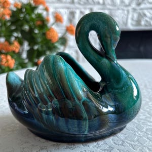 May include: A ceramic swan ornament with a glossy, teal and green glaze. The swan has a curved neck and detailed wing feathers. The ornament is a decorative item, likely for display.