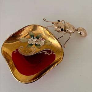 May include: A gold-colored metal ant figurine perched on the edge of a small, square, gold-rimmed dish. The dish has a red interior and is decorated with painted flowers in shades of white, green, and pink.