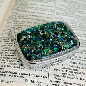 May include: A rectangular silver-toned metal pill box featuring a lid inlaid with a mosaic of crushed iridescent abalone shell fragments in shades of deep blue, emerald green, and white, set within a smooth, polished metallic frame.