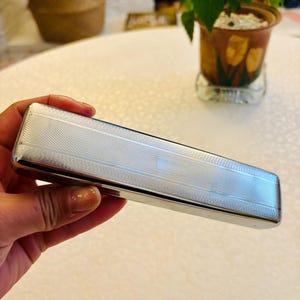 May include: A rectangular silver-toned metal case with a textured cross-hatch pattern on the lid and a smooth, blank rectangular panel in the centre. The case features rounded edges and a polished finish, presenting a sleek and uniform appearance.