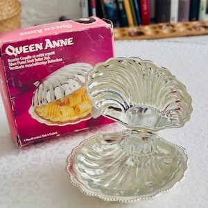 May include: A silver-plated butter dish shaped like an open seashell with a hinged lid and a beaded edge. The dish is displayed next to its original pink cardboard box, which features the text Queen Anne and Silver Plated Shell Butter Dish.