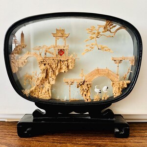 May include: An oval-shaped cork carving display encased in a black frame with a matching black stand. The intricate light brown cork sculpture depicts a landscape with pagodas, arched bridges, rocky cliffs, pine trees, and two small white cranes.