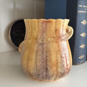 May include: A ceramic vase with a textured surface and a warm color palette of yellow, brown, and orange. The vase has a ruffled rim and two small handles. The vase is displayed on a white shelf with books in the background.