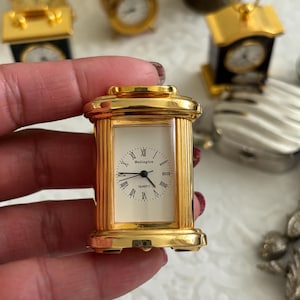 May include: A gold-colored, rectangular quartz clock with a white face and black Roman numerals. The clock face reads "Wellington" and "Quartz". The clock is held in a hand, with other clocks in the background.