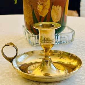 May include: A polished brass chamberstick candle holder featuring a wide, circular drip tray base, a central cylindrical socket for a candle, and a single, rounded finger loop handle attached to the side of the tray for easy carrying.