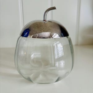 May include: A clear glass jar with a silver lid and stem-like handle. The jar has a rounded shape, and the lid is shaped like an apple. The jar is likely used for storing sugar or other small items.